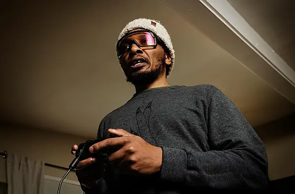 Del is standing with a video game controller in his hand. He is wearing a dark grey long-sleeve shirt, a grey beanie cap and a large pair of polarized goggles that wrap around his face. He is looking intensely at what can be assumed is the TV screen.
