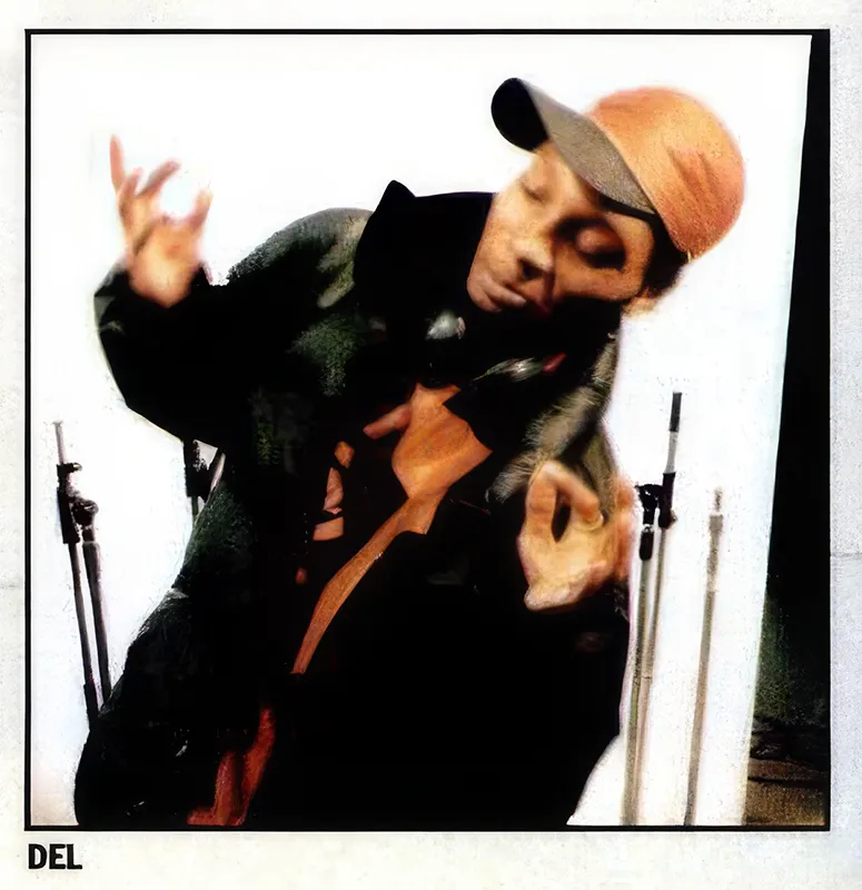 Del the Funky Homosapien is drissed in a black winter coat, with an orange baseball cap. He is making a silly pose leaning to the side, one hand open to the sky, and another making a pinching gesture. Recording mic-stands can be seen in the background. The photograph was taken in a recording studio.