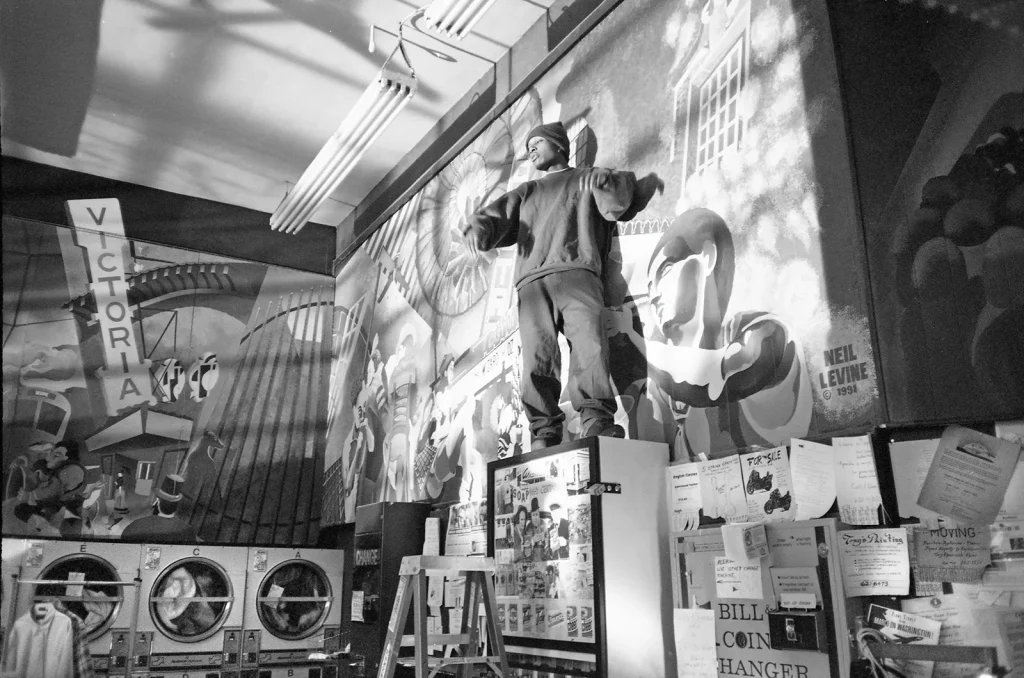 Del the Funky Homosapien stands atop a soap dispenser in the Little Hollywood Launderette in San Francisco, CA. The photograph is taken at angle and from below, looking up at Del. It is shot in black and white with a wide angle lens. A large mural of San Francisco's castro District appears behind Del who has his arms raised as he rhymes to the music.