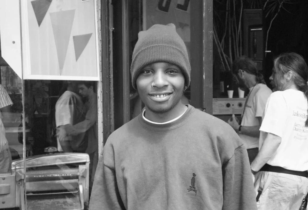 Del the Funky Homosapien appears outside of the Little Hollywood Launderette in San Francisco, CA. The photograph is in black and white and shot by owner & operator of the laundromat, Dave Glass. Production crew can be seen in the background, presumedly preparing for filming. Del is smiling directly at the camera wearing a beanie cap and sweater and sporting a nose ring.