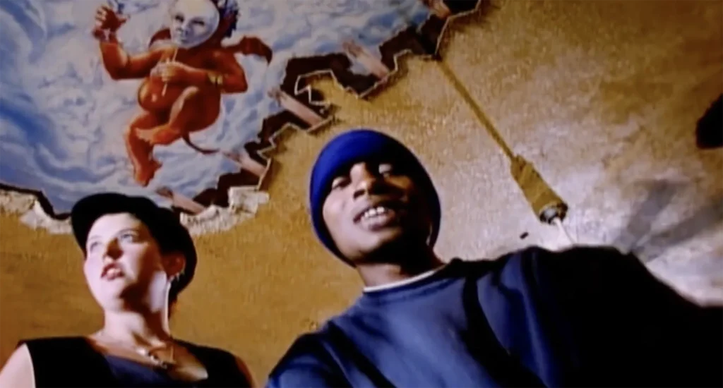 Del the Funky Homosapien looks down at the camera, which is positioned low. A white woman with short hair and many earrings, wearing a newsboy cap, stands next to him, staring off into the distance. A mural appears above their heads, depicting a devil-like cherub holding a Commedia dell’arte mask.