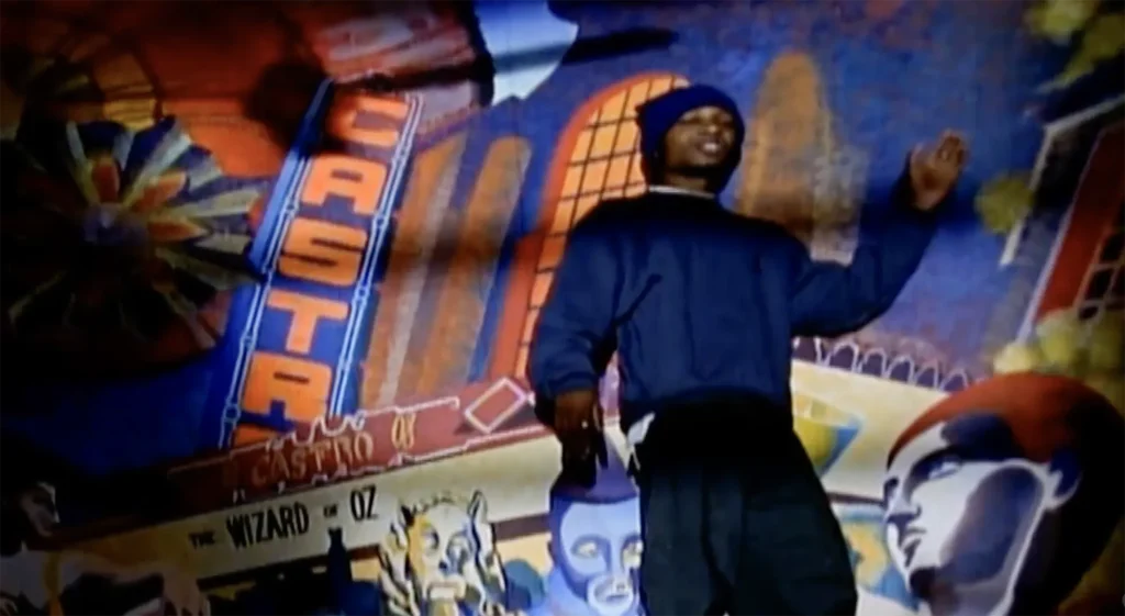 Del the Funky Homosapien performs “Made in America” in front of a large Castro District mural inside the Little Hollywood Launderette in San Francisco, CA. Del has one arm raised and appears lively.