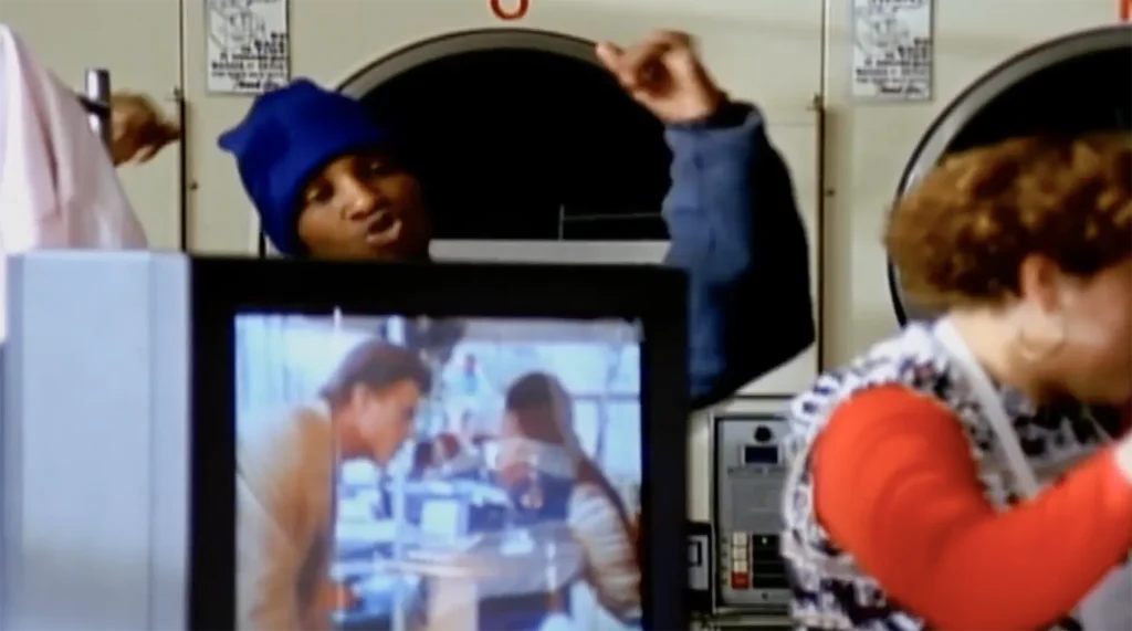 Del the Funky Homosapien performs “Made in America” in front of a set of laundry machines, his arms raised and fists clenched. Out of focus in the foreground, a woman appears to be folding clothes. A television beside her plays scenes from the movie of the same name, featuring images of Ted Danson.