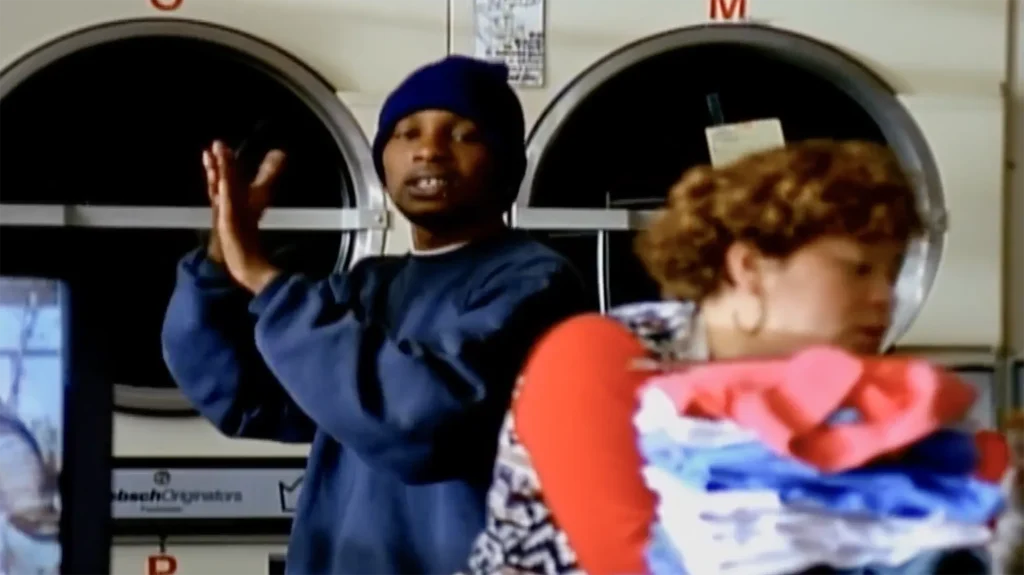 Del the Funky Homosapien performs “Made in America” in front of a set of laundry machines, his hands clasped. Out of focus in the foreground, a woman appears to be folding clothes.