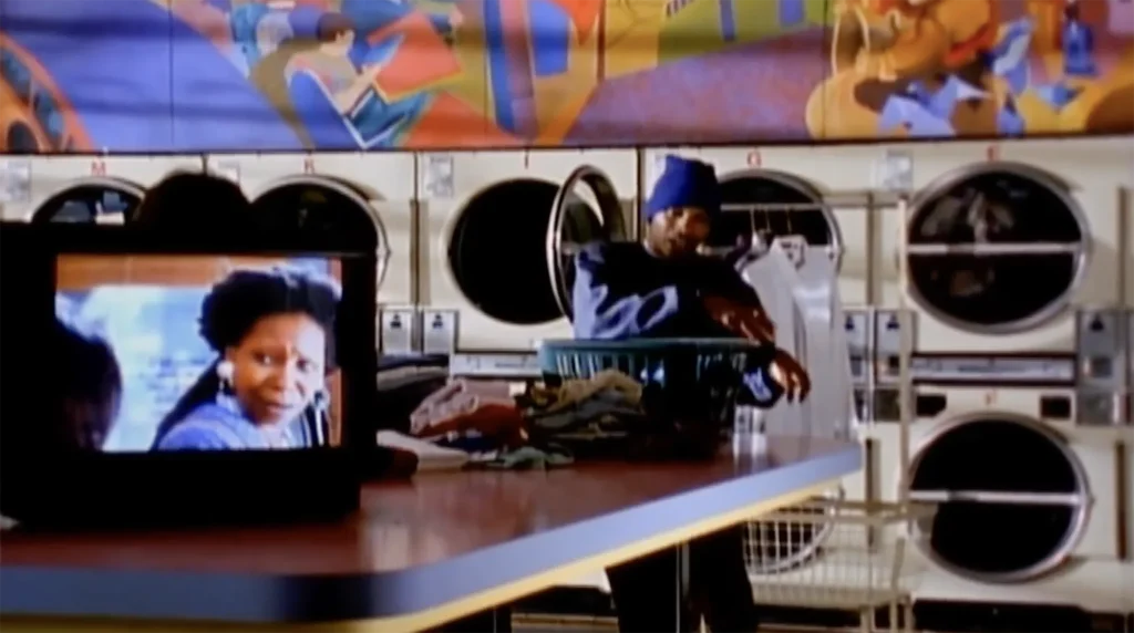 Del the Funky Homosapien appears in a long telephoto shot, his arms resting on a basket of clothes as washing machines tumble behind him. In the foreground, a television rests on a table, projecting a scene from the movie "Made in America" featuring Whoopi Goldberg.