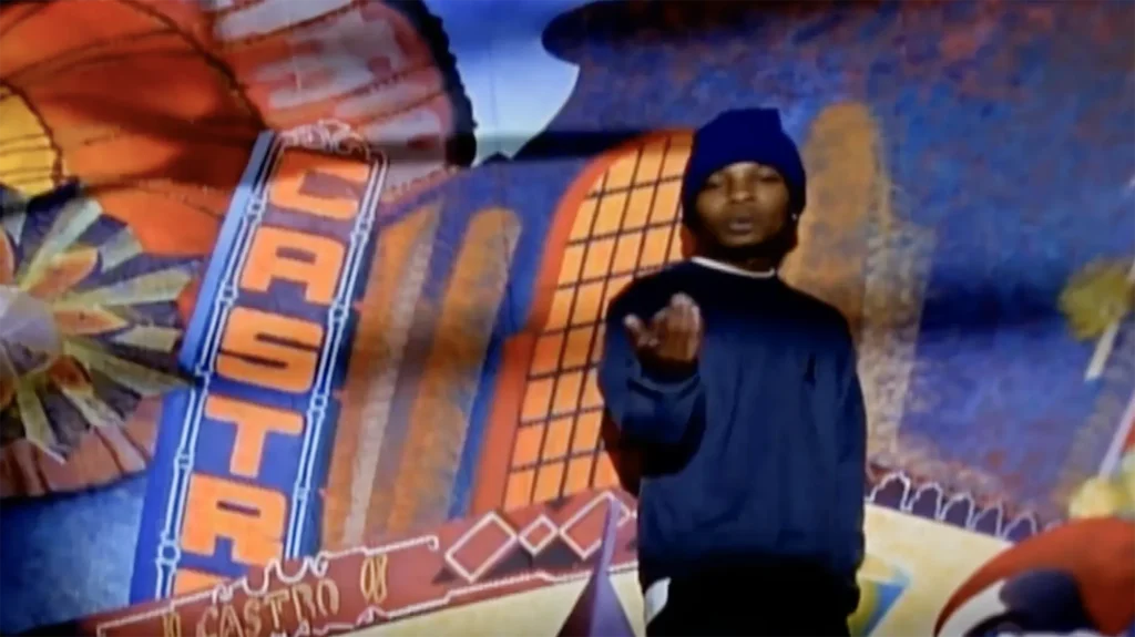 Del the Funky Homosapien stands in front of a large Castro District mural inside the Little Hollywood Launderette. He is wearing a blue beanie and a blue sweatshirt, with his hand extended.