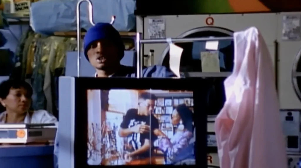 Del the Funky Homosapien appears through a drying rack, performing his song “Made in America.” A woman, possibly an employee of the Little Hollywood Launderette’s dry-and-fold service, is sitting behind a table with a scale in front of her. Her face appears amused, her mouth closed and head tilted.