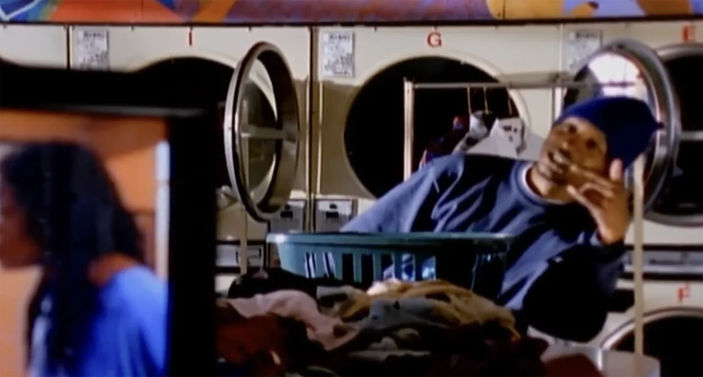 Del the Funky Homosapien stands in front of a set of laundry machines and a hanging rack, pointing towards the camera. A television screen, displaying scenes from the movie "Made in America," plays slightly out of focus in the foreground.