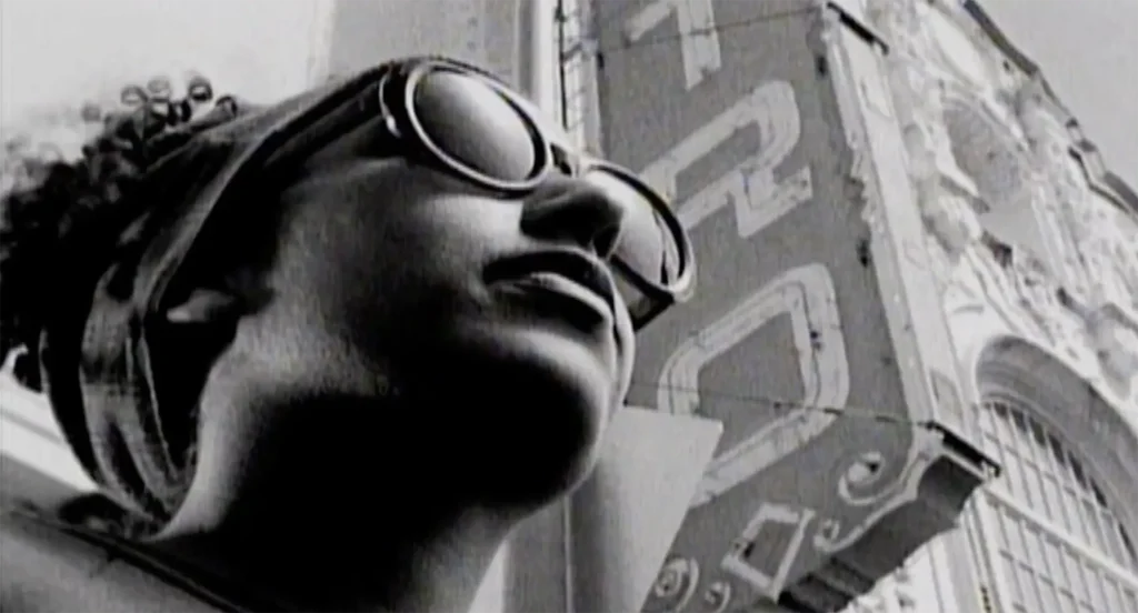 A woman wearing sunglasses stares into the distance as the camera, positioned low, looks up at her. The Castro Theater marquee looms large above her. This is a screenshot from the 1993 Del the Funky Homosapien music video, “Made in America.”