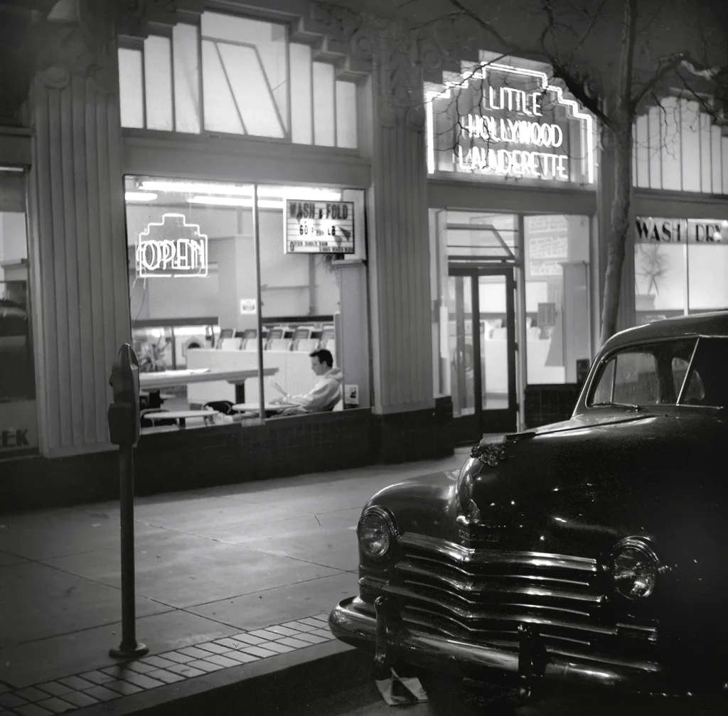 The Little Hollywood Launderette is lit up at night in this black-and-white photograph taken from outside. Large windows frame the bustling indoor space, flanked by impressive glass arches above them. A large neon marquee displays the laundromat’s name above the door. In another window, a neon sign glows with the word “Open.” A classic car, possibly from the 1940s or 1950s, is parked out front on Market Street, characterized by its rounded design, prominent chrome grille, and hood ornament. Photograph by Dave Glass.