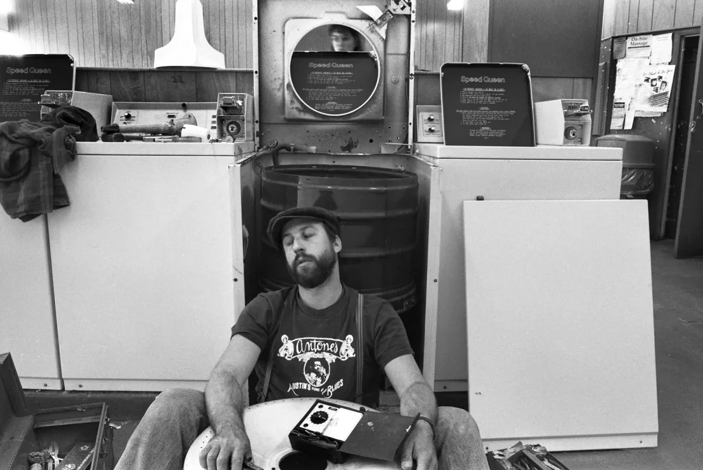 Dave Glass sits on the floor among a set of washing machines, one of which is largely disassembled for repair. He straddles a large metallic drum, clearly a part of the machine he is working on. An open toolbox rests near his leg. Dave is wearing a t-shirt with suspenders and a newsboy cap. He has a full beard, and his eyes are closed, with his head slightly tilted, suggesting the weight of a heavy workload ahead. The reflection of an unknown photographer is visible in a reflective part of the open washing machine. The photograph is undated.