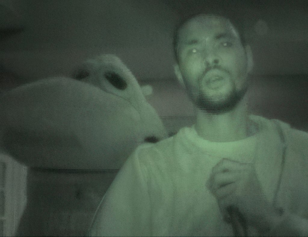 Yameen & Tajai perform on stage as The Entity & SupremeEx. The photo is taken from within the crowd and shot using night-vision technology, giving the image an uncanny green glow. Fittingly, Tajai’s eyes shine inhumanly as a result of the effect. SupremeEx wears a large, oversized costume head of indiscernible origin.